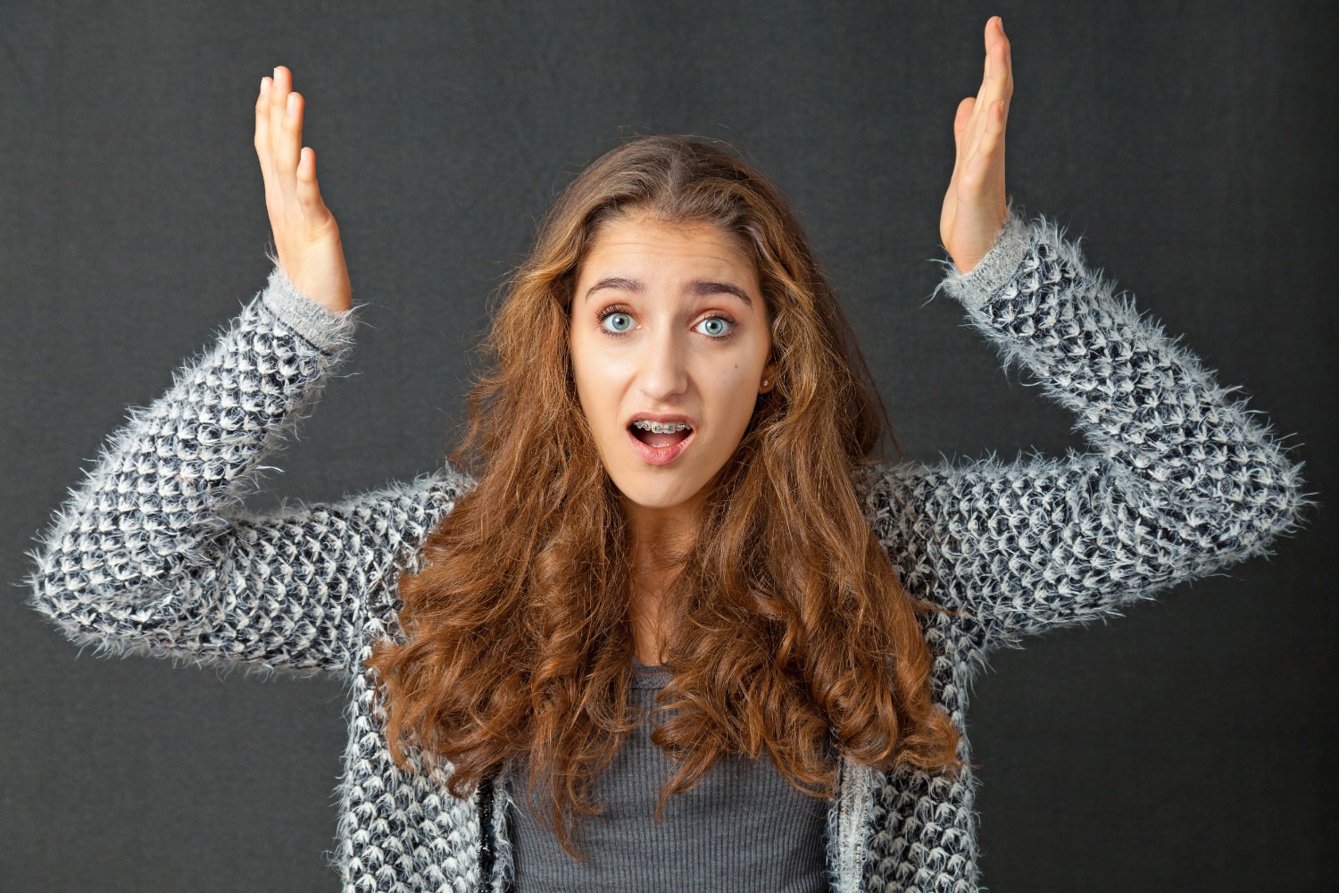 A person with long hair and a patterned sweater raises both hands, appearing surprised, against a plain dark background. No landmarks visible.