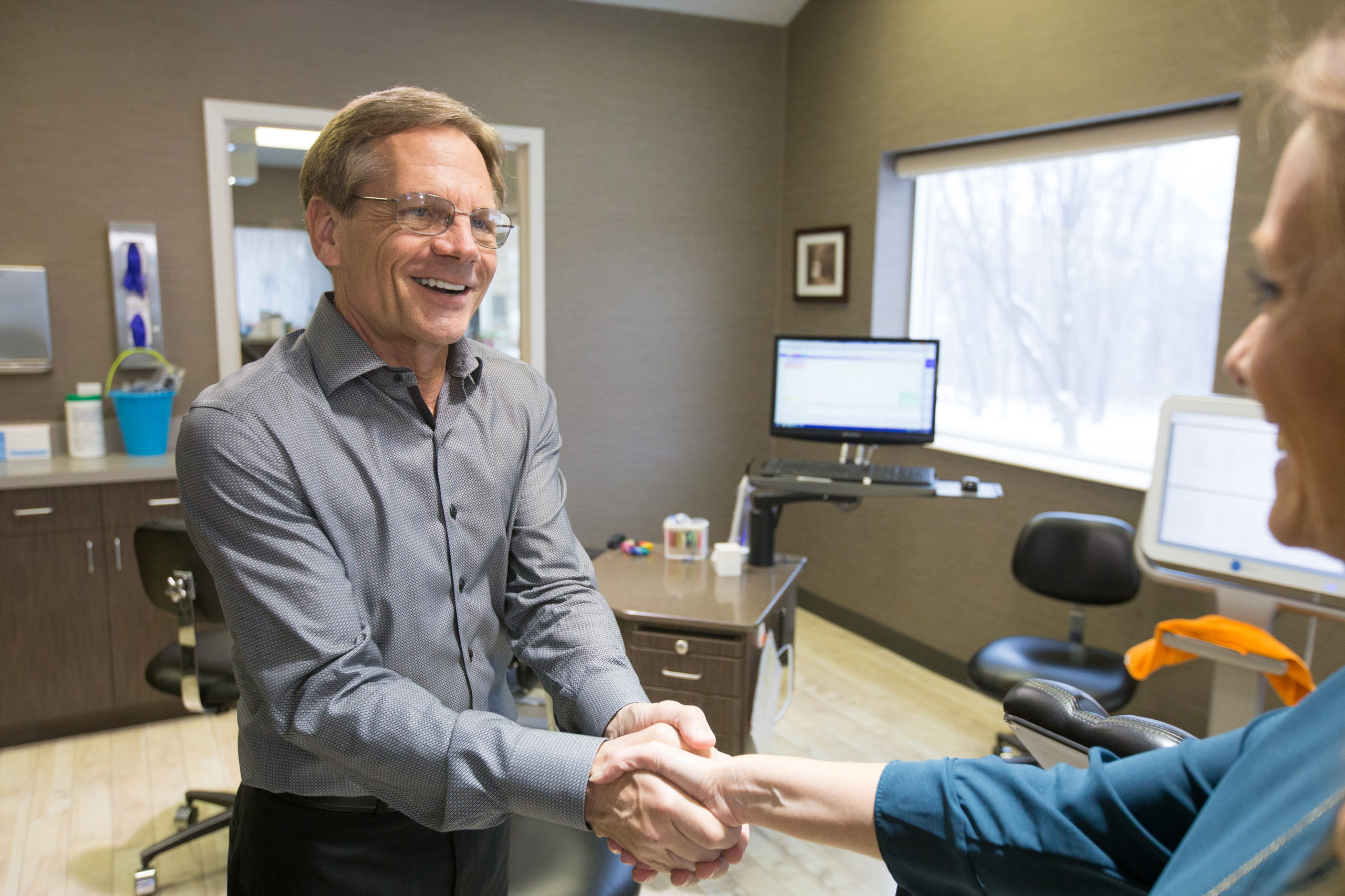Two people shake hands in a modern office. One smiles warmly. A window shows a snowy scene outside. Office equipment is visible.
