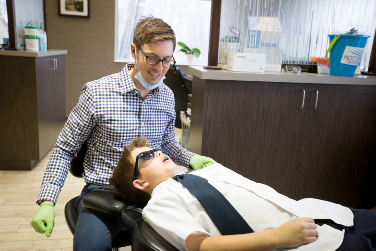 A person wearing gloves attends to a child reclining in a dental chair, inside a modern dental office with various equipment visible.