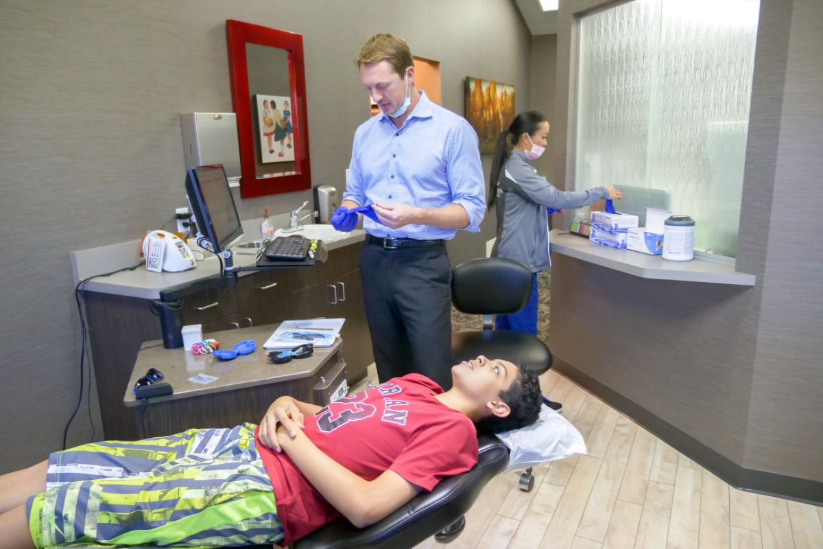 A person in a dental office prepares tools while another rests on the chair. A third person organizes supplies in the background.