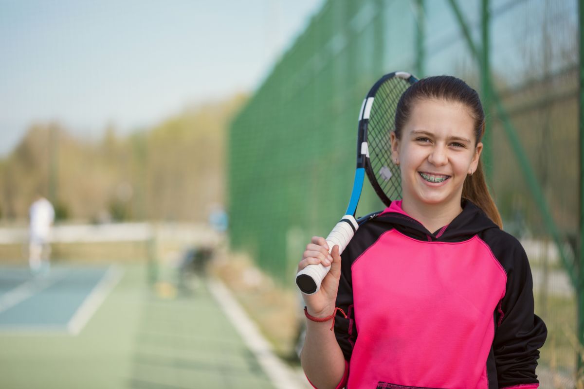 A young person with a tennis racket smiles near a tennis court. Another person is in the background, with trees along the fence.