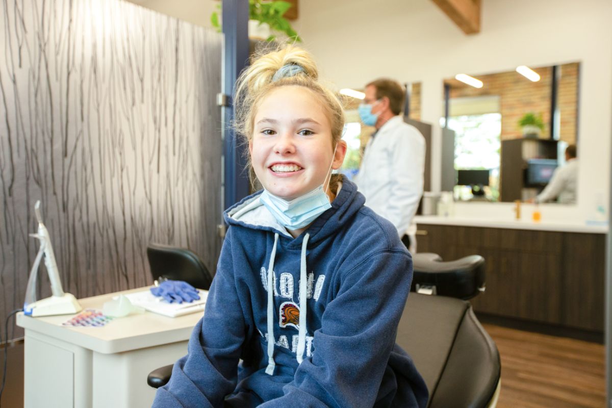 Young person smiling in a dental office, wearing a hoodie. Mask is around neck. Person in background is wearing a white coat and mask.