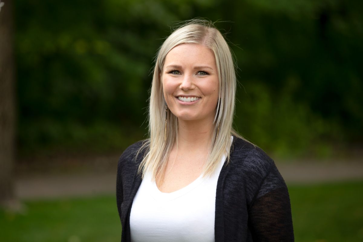 A person with long, blonde hair smiles outdoors. Green foliage and a pathway are visible in the blurred background. No landmarks present.