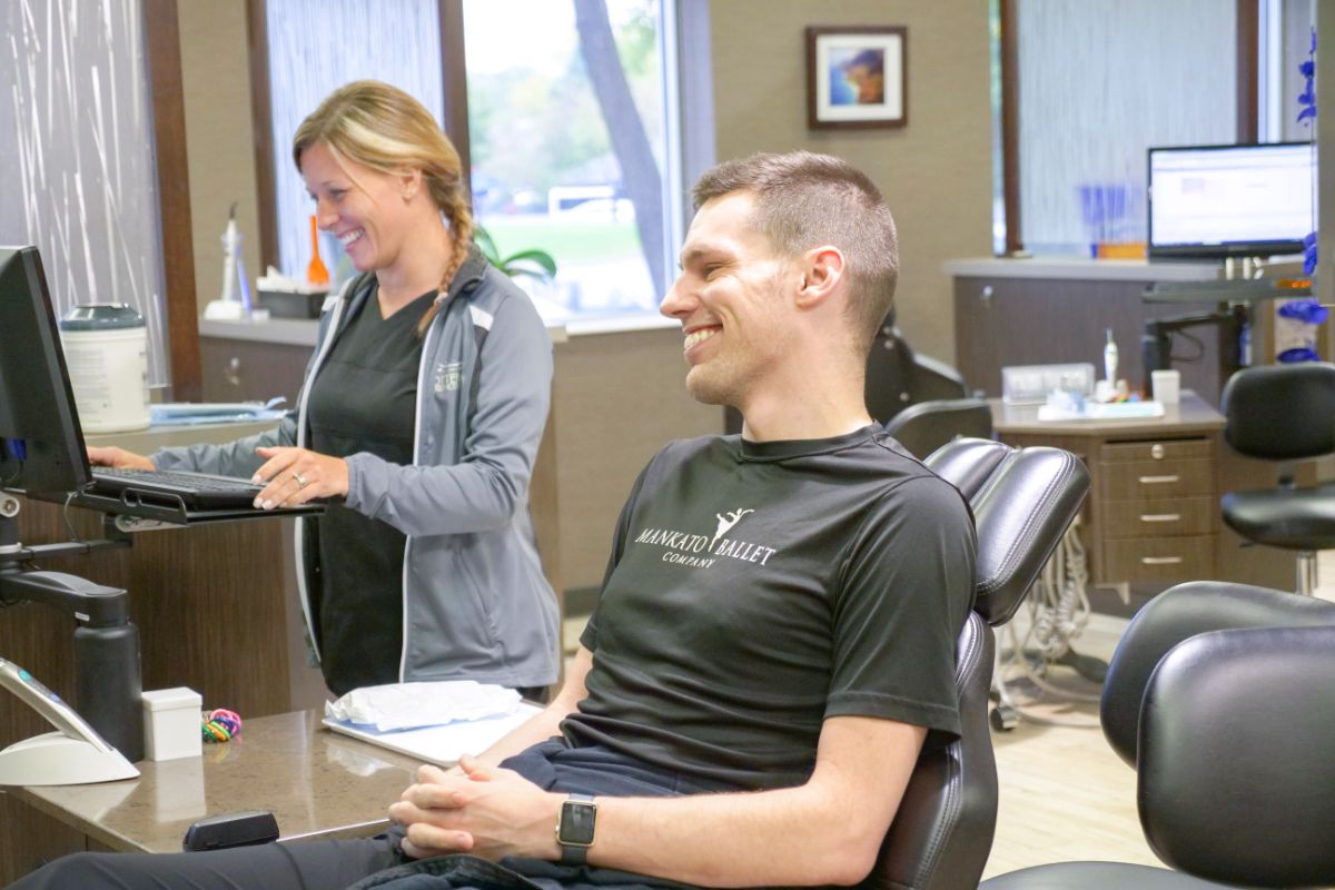 A person sits in a dental chair smiling, while another person in uniform works at a computer in a modern dental office.