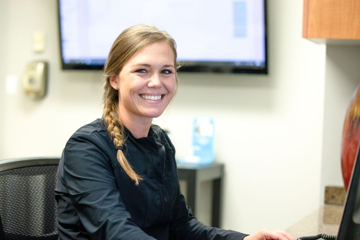 A person with a braid smiles at a desk in an office setting, with a monitor in the background and a cabinet nearby.