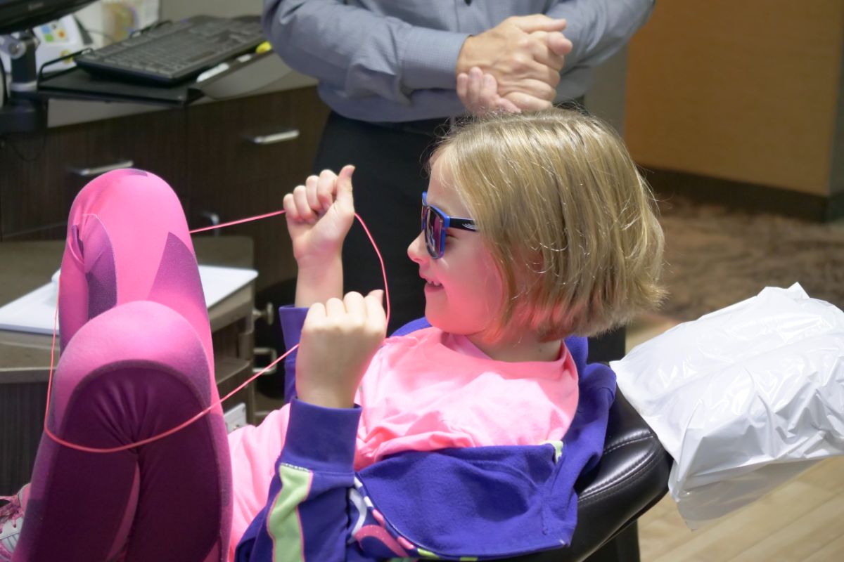 A child wearing sunglasses sits on a chair playing with a pink string, while a person stands nearby in an indoor setting.