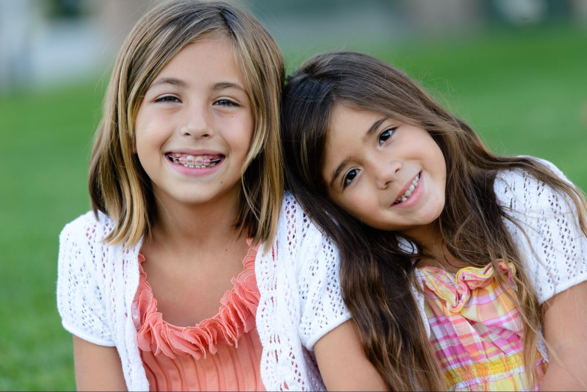 Two children smiling, sitting close together on grass, wearing white cardigans and colorful dresses. Blurred background enhances their joyful expressions.