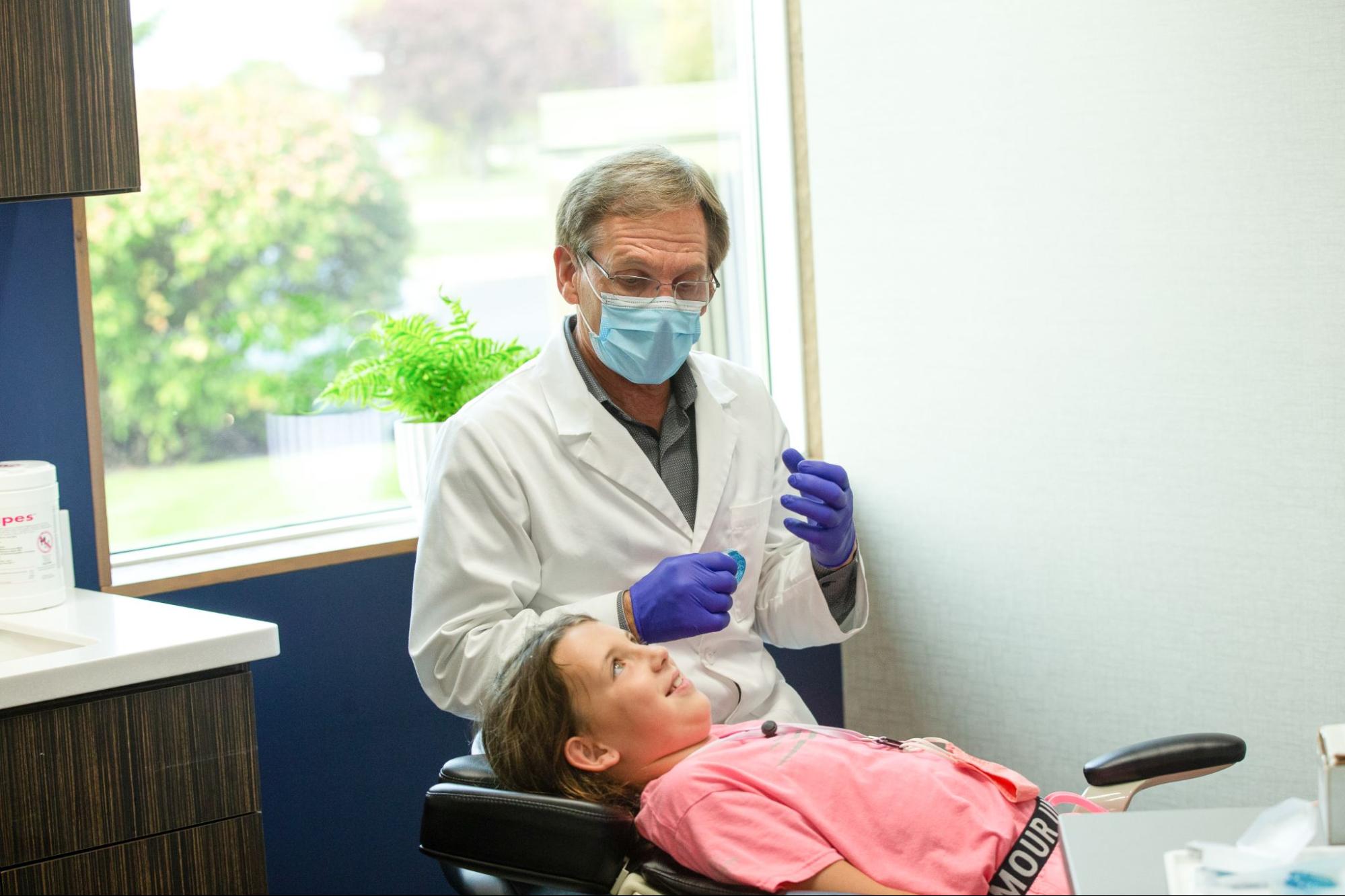 A person in a dental office, wearing a mask and gloves, talks to a child in the dental chair near a bright window.