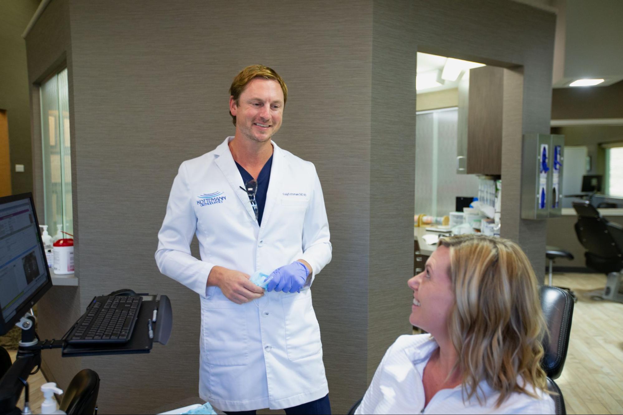A person in a dental office, wearing a white coat and gloves, stands by a seated person, holding dental supplies. Office equipment visible.