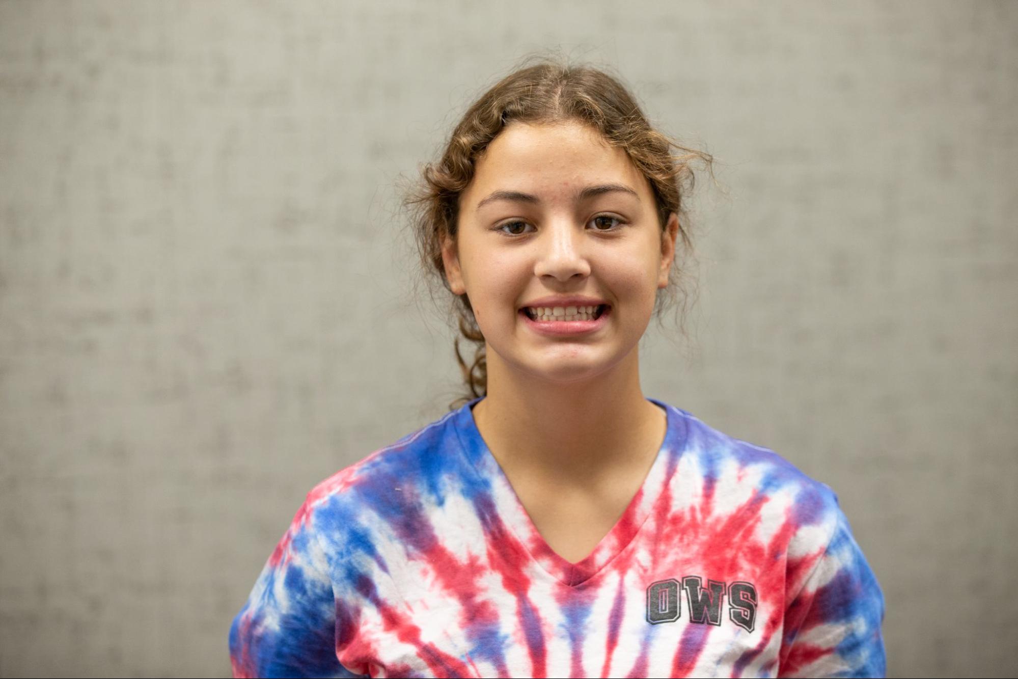 A smiling person wears a colorful tie-dye shirt with "OWS" letters, standing against a neutral, textured background.