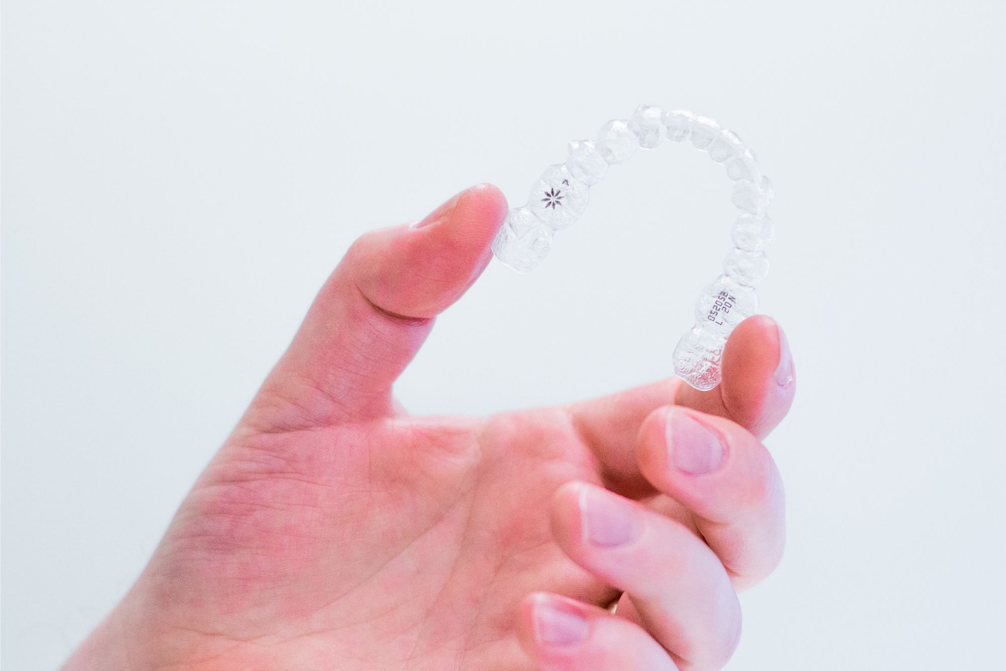 A person's hand holds a clear dental aligner against a plain white background, showcasing the aligner's translucent plastic design and intricate details.
