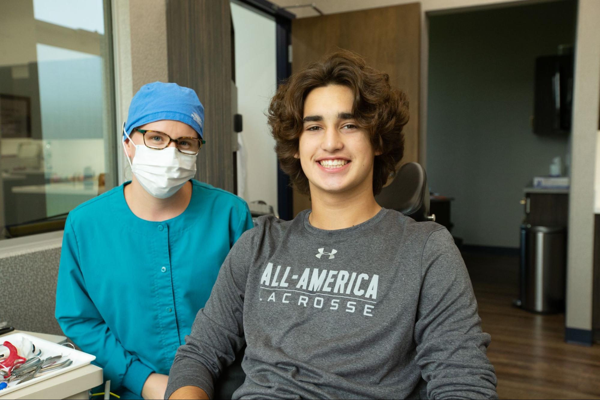 A person wearing scrubs and a mask stands next to a smiling young person in a dental office setting, with dental tools visible.