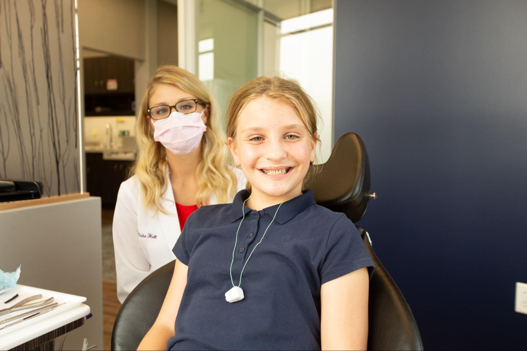 A child sits in a dental chair, smiling at the camera. A person wearing a mask and white coat stands behind in a dental office.