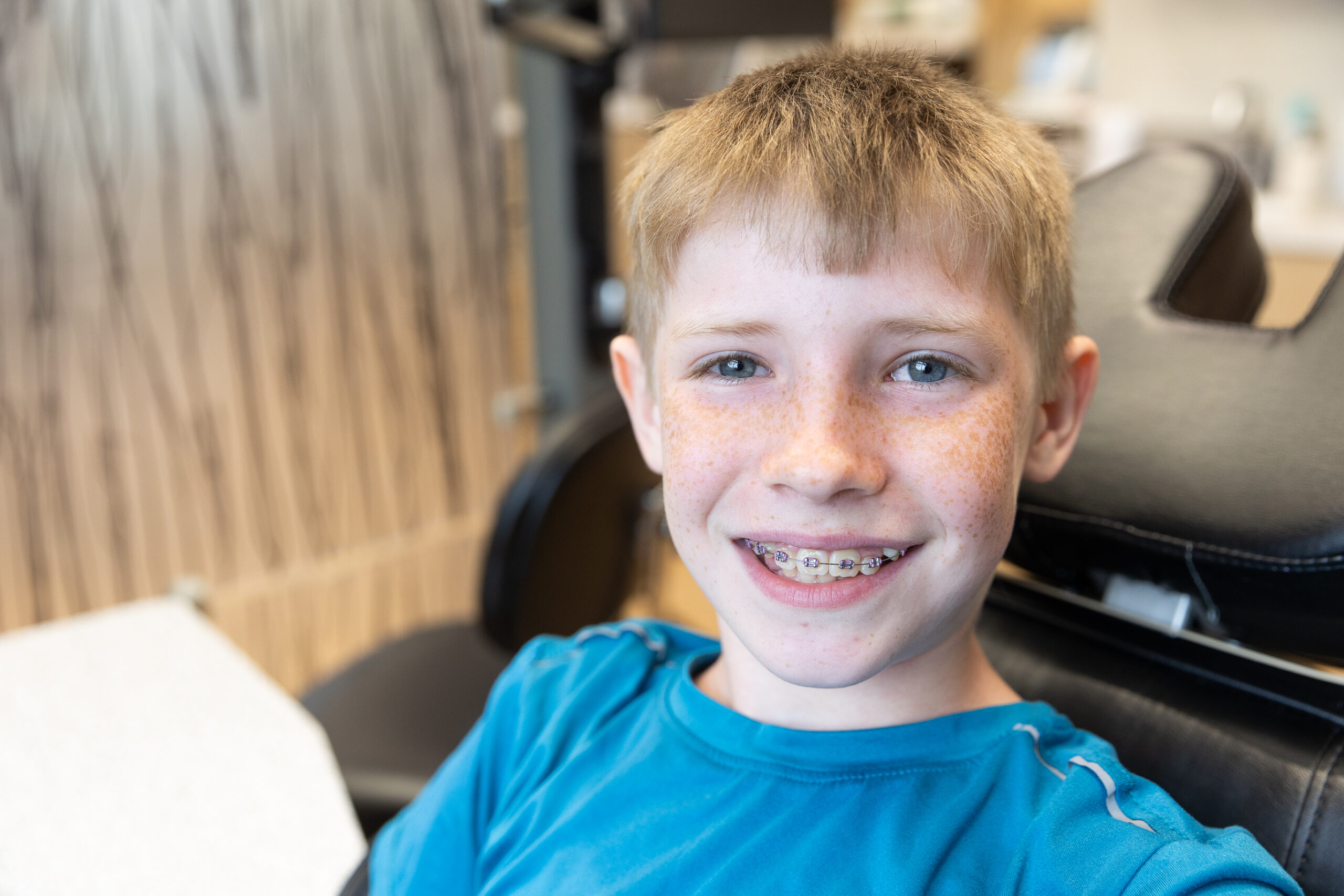 A child wearing braces smiles while seated in a dental office, with modern equipment and decor visible in the background.