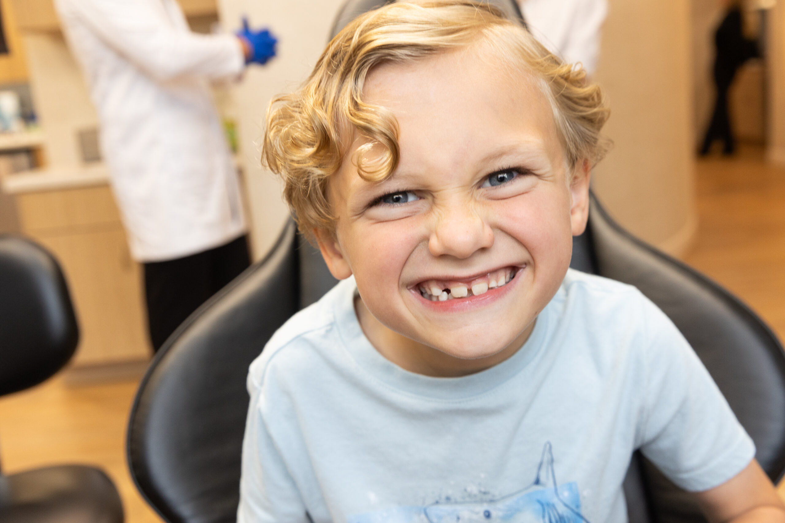Smiling child with curly hair sits in a dental chair. Background shows two persons in white coats attending to dental tasks.