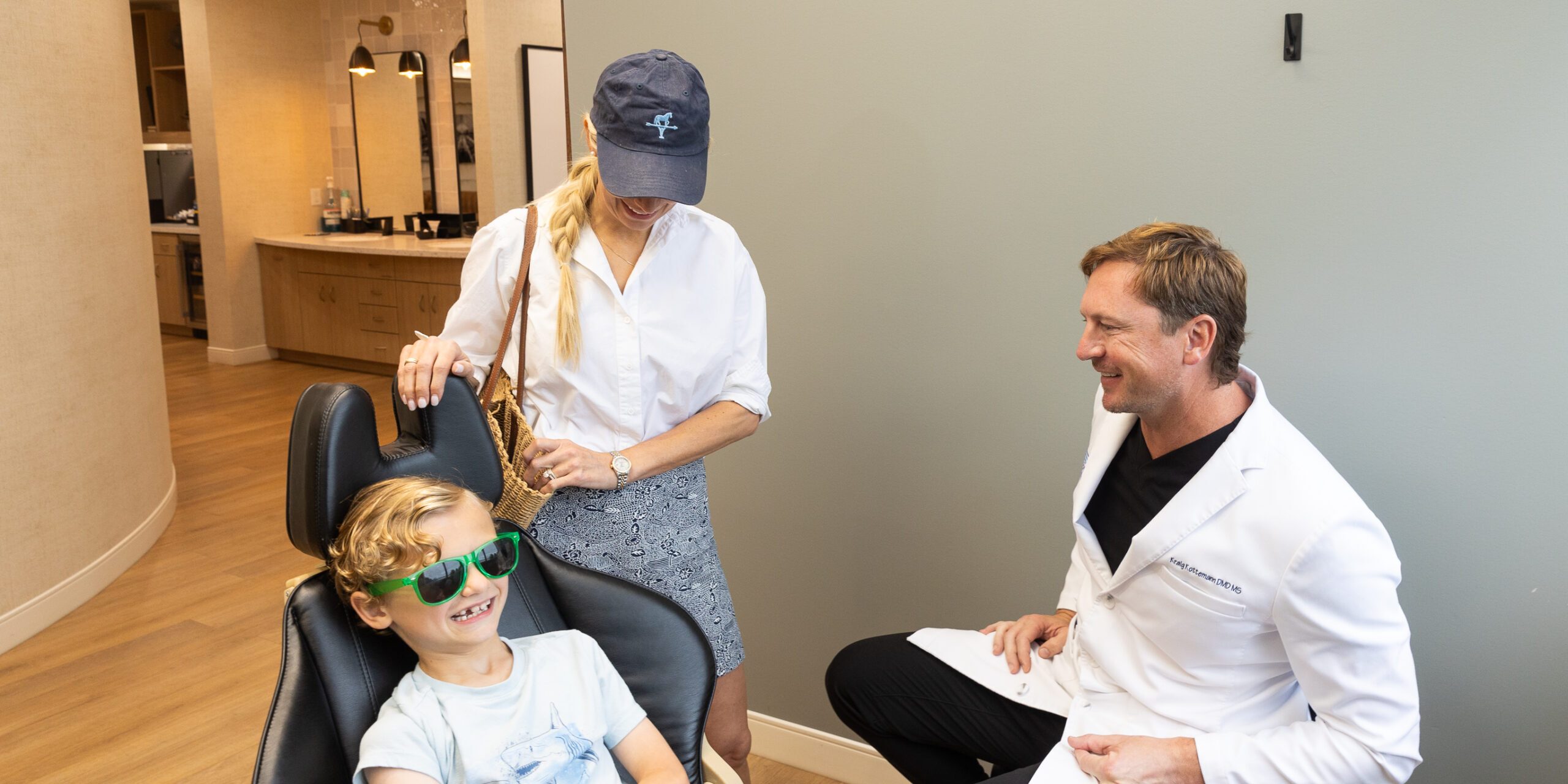 A person in a white coat chats with a seated child wearing sunglasses, while another person stands beside them in a modern office.
