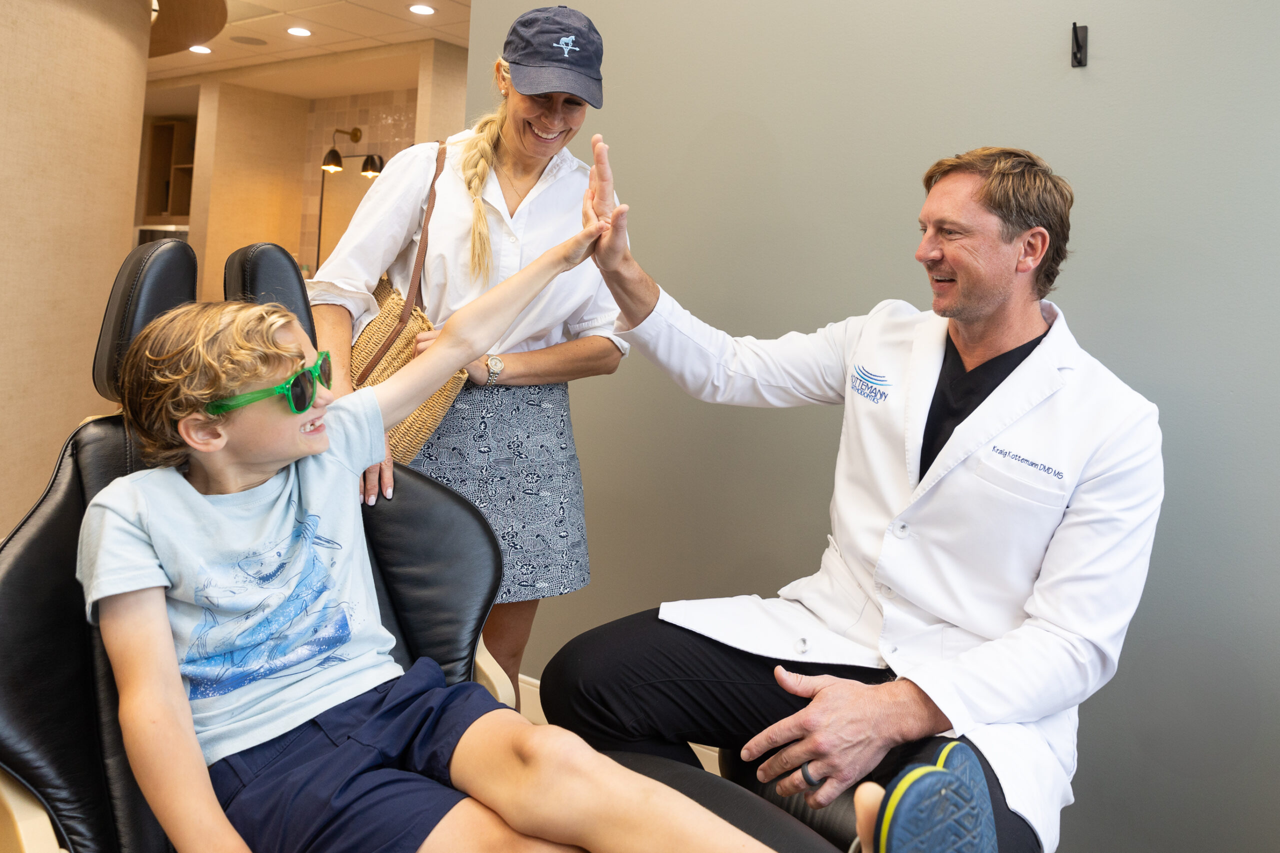 A child wearing green sunglasses high-fives a person in a white coat, while another person looks on, smiling, in a medical office.