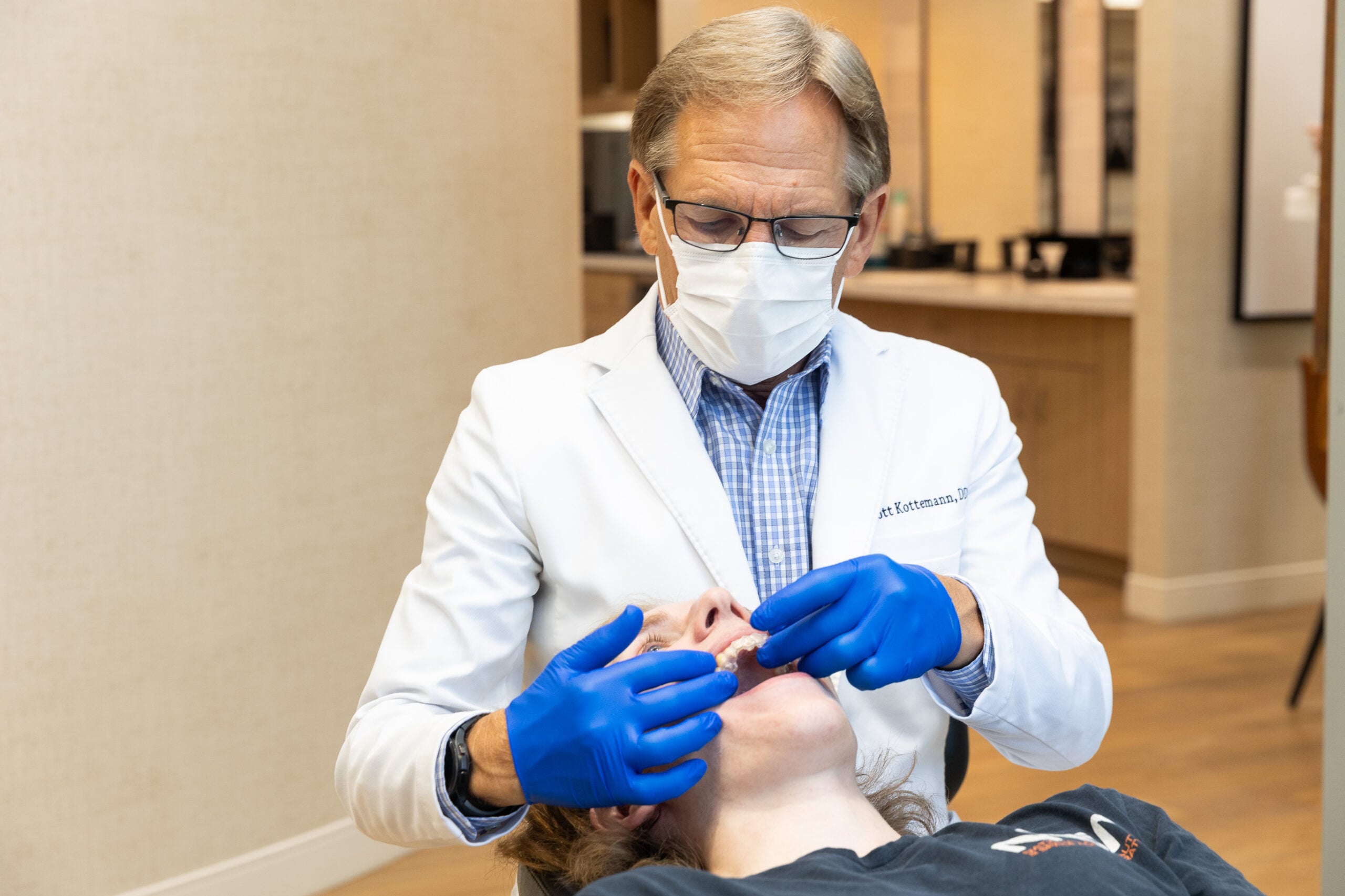 A person in a white coat and gloves examines another person's mouth in a clinical setting. Books and mirrors are visible in the background.