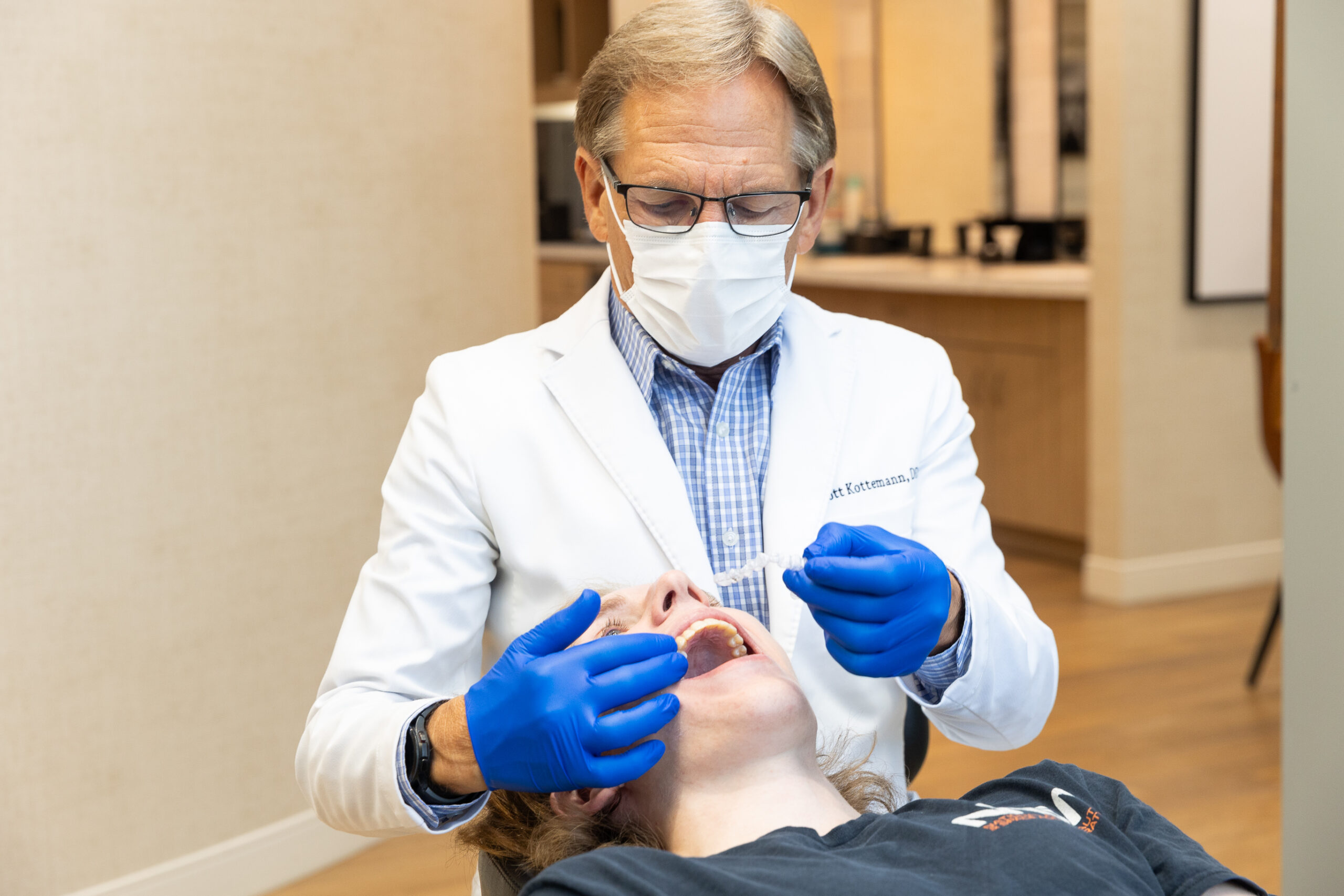 A person in a white coat and blue gloves examines another person's teeth in a dental office setting. The person is wearing a mask.
