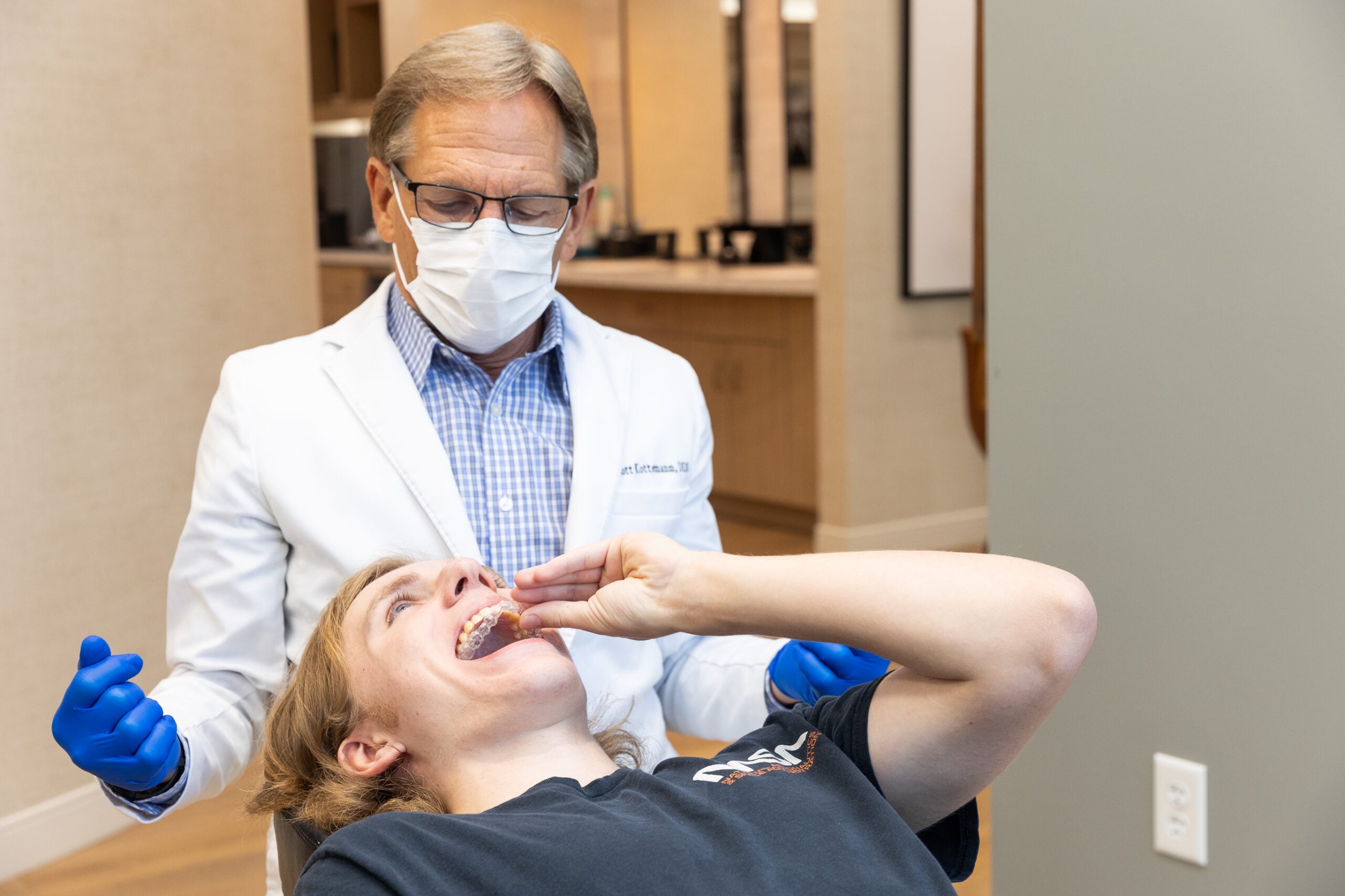 A person wearing a lab coat and mask examines another person lying back with their mouth open, possibly in a dental or medical setting.