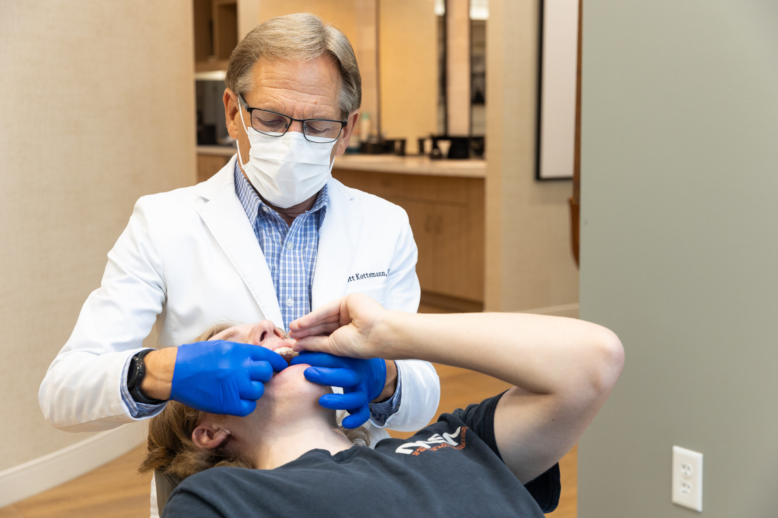 A person in a white coat examines another person's mouth, wearing gloves and a mask, in a modern dental office setting.
