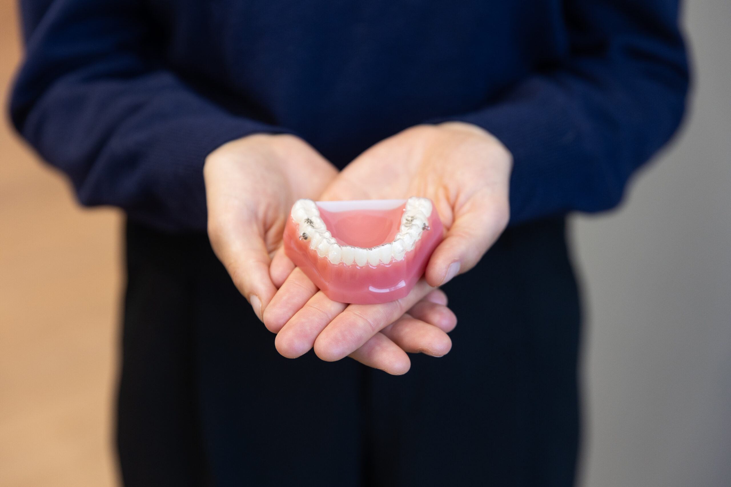 A person holds a dental model with a set of teeth, wearing a dark sweater. The background is out of focus.