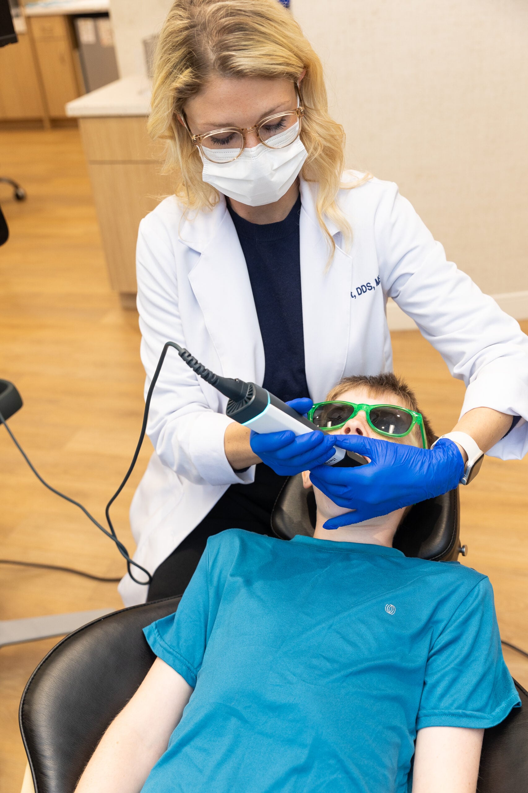 A person in a dental office inspects a patient's teeth. The patient wears green sunglasses, while the person wears a white coat and blue gloves.