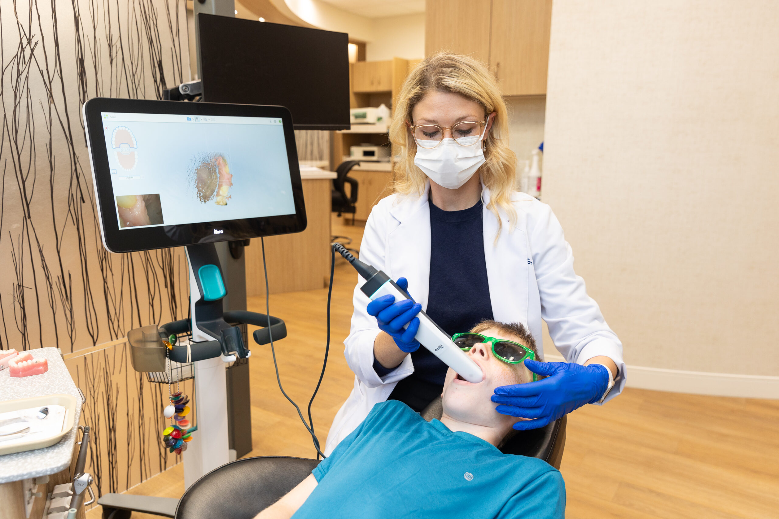 A person is performing a dental procedure on another person in a clinic, using equipment. Both wear masks; one also wears gloves and glasses.