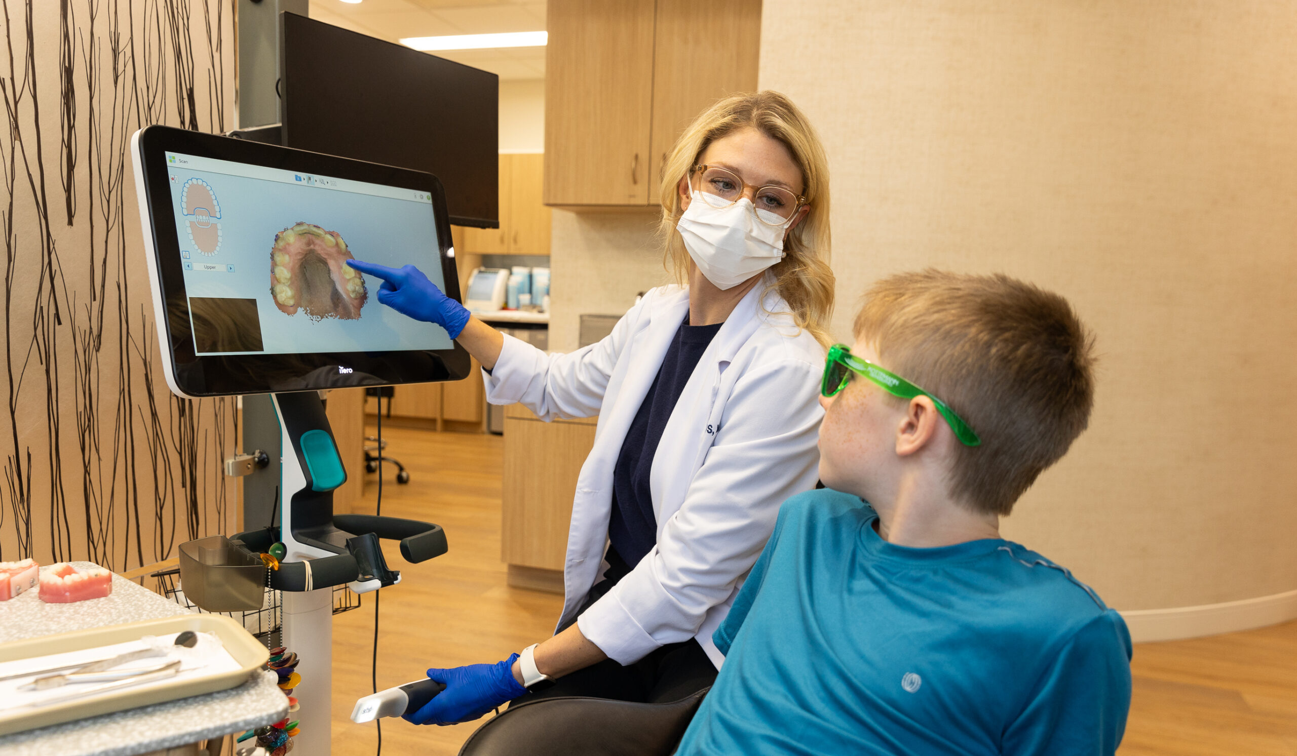 A dental professional explains a 3D teeth scan to a child wearing protective glasses in a modern dental office setting.