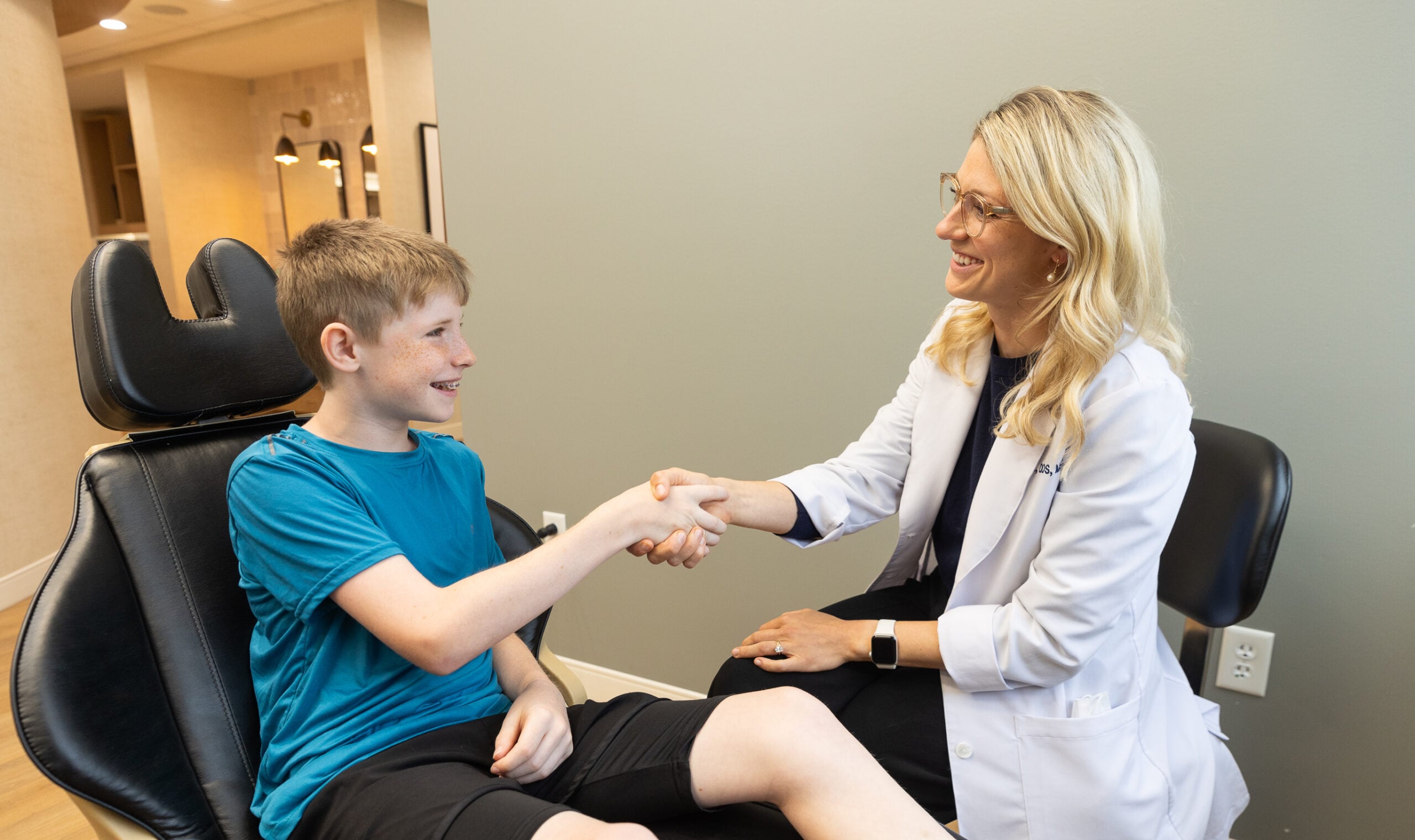 A person in a white coat shakes hands with a smiling child sitting in a dental chair, inside a well-lit room.