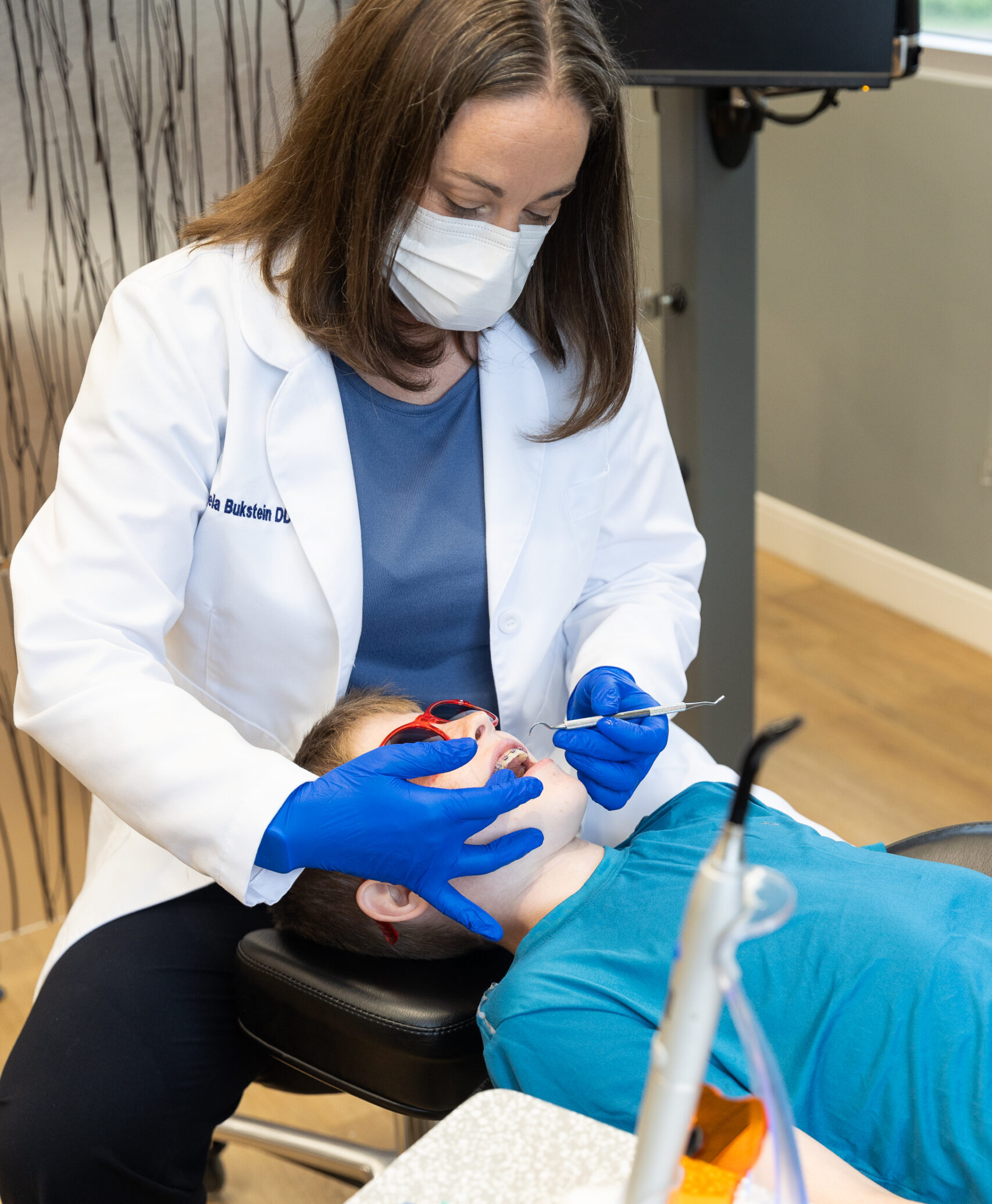 A person in a dental office examines a reclined child wearing sunglasses. Dental tools and colorful hairbands are visible in the foreground.