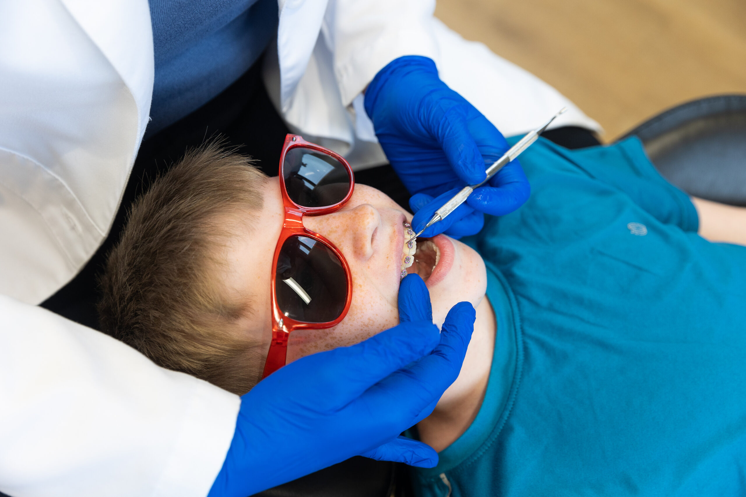A person wearing blue gloves examines a child's braces. The child wears a teal shirt and red sunglasses, reclining in a dental chair.