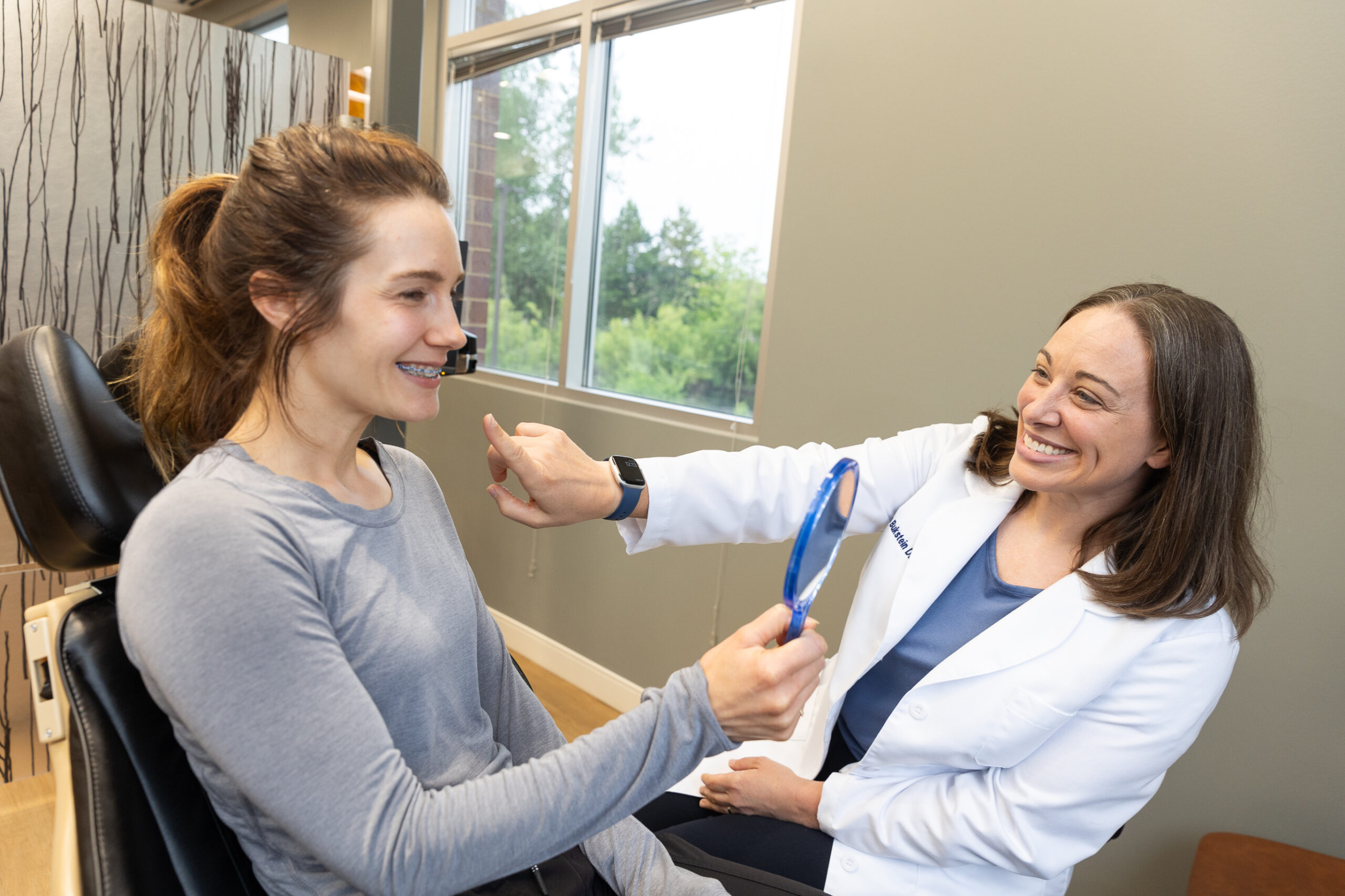 Two people in a medical office. One sits holding a mirror while the other, in a white coat, gestures and smiles.