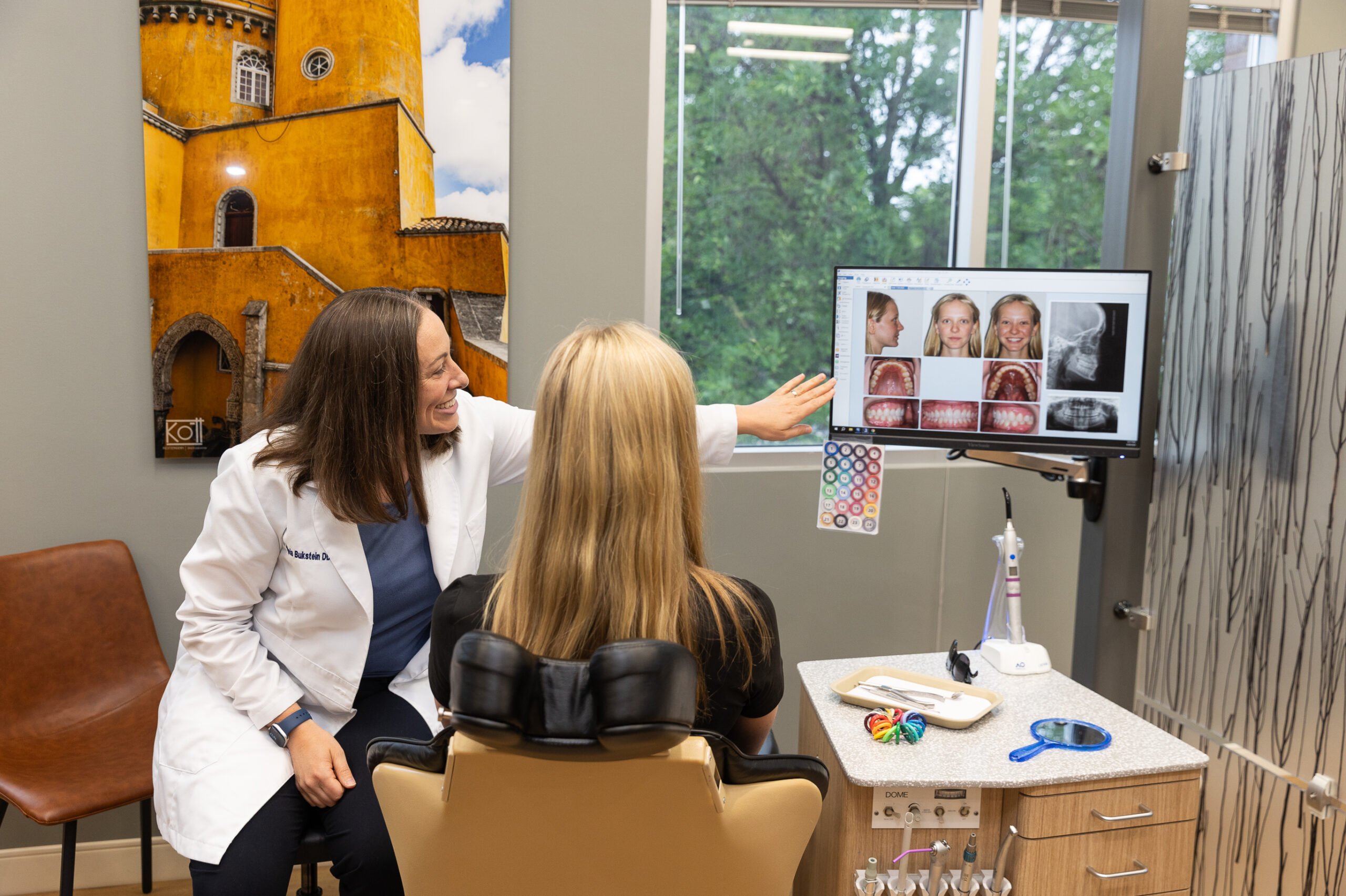 A person in a white coat explains dental images to another person in an office setting, with a colorful building photo on the wall.