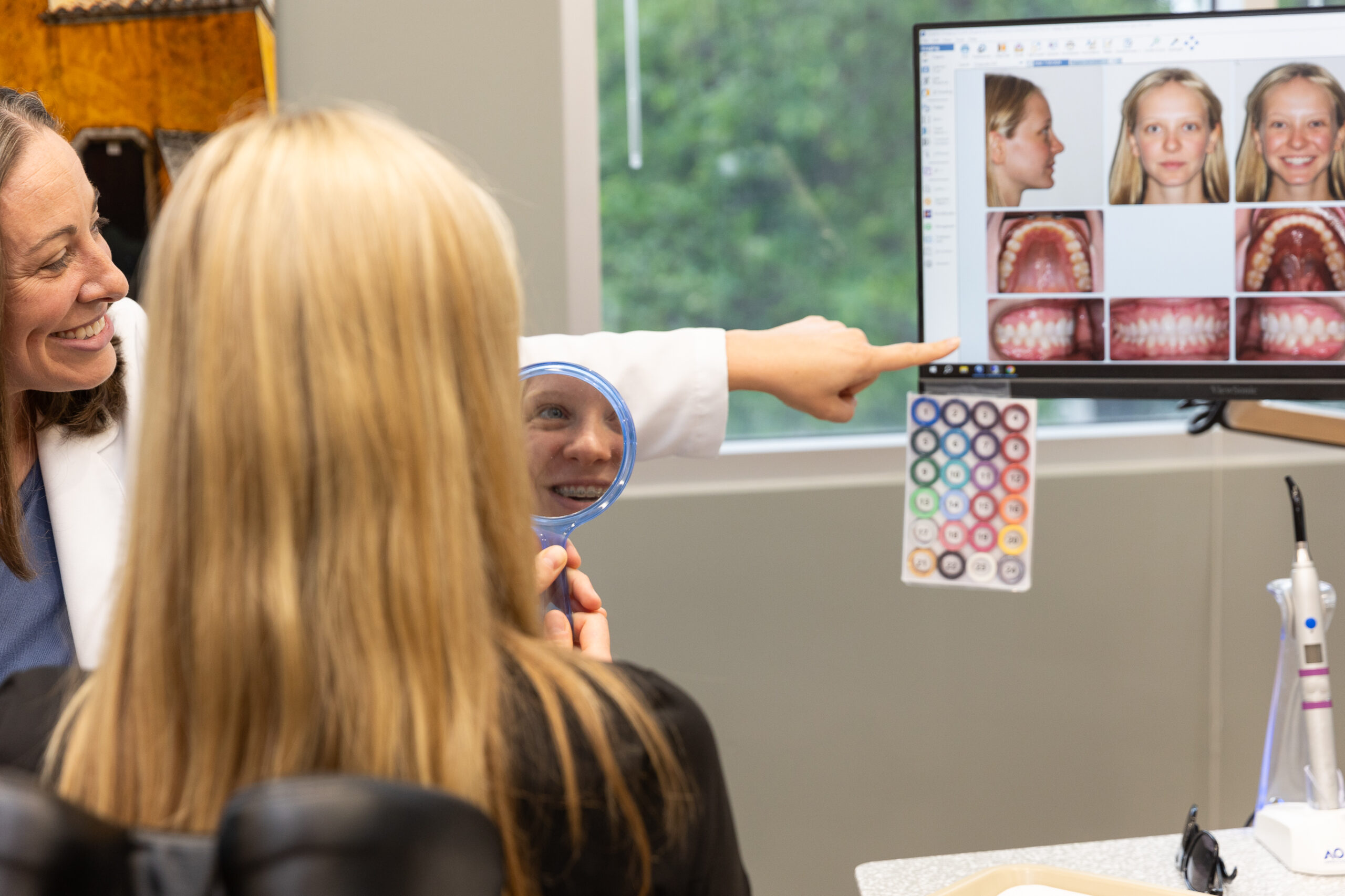 A person shows another a dental image on a computer. The seated person holds a mirror, reflecting their smile.