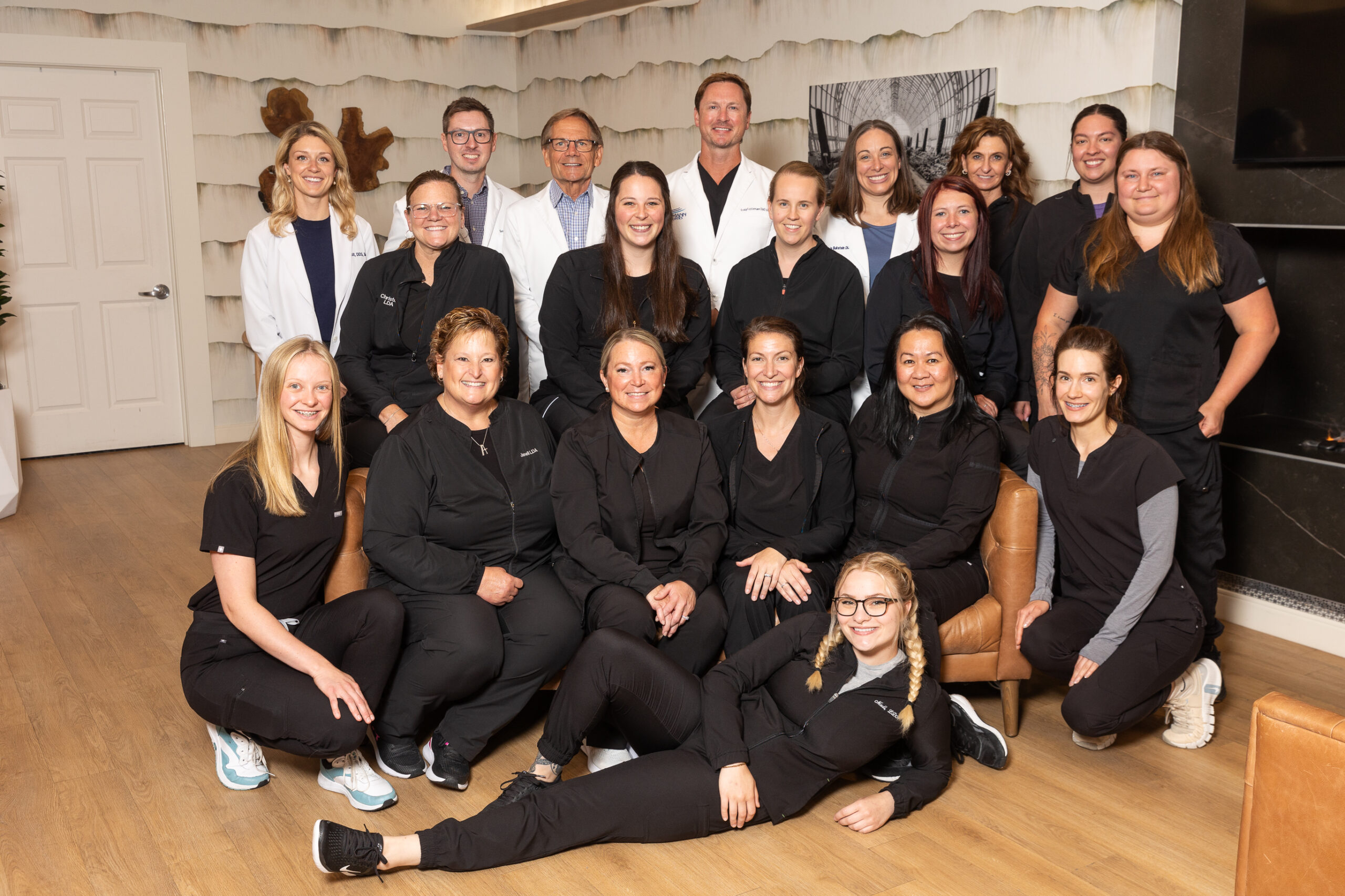 A group of seventeen people, wearing black and white attire, pose indoors on a wooden floor with light-colored textured walls in the background.