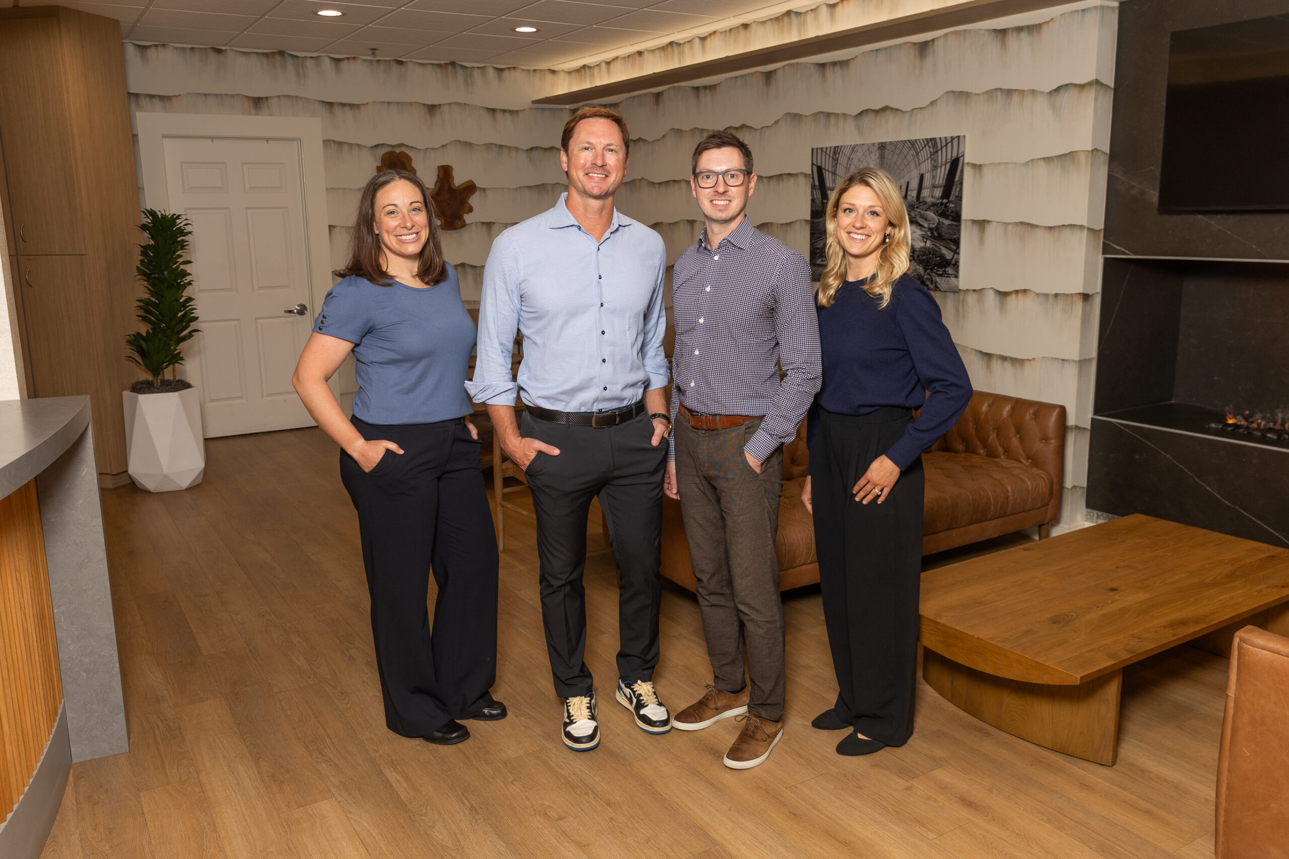 Four people stand smiling in a modern, cozy office with wooden flooring, a sofa, and artistic decor. There's no recognizable landmark present.